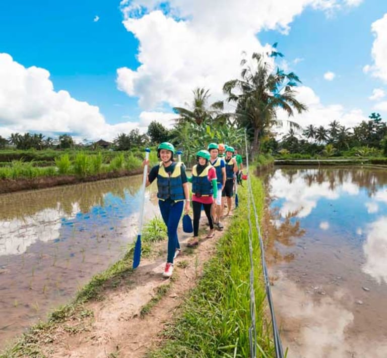 A group of tourists wearing life jackets and helmets walking along a path in Bali rice fields.