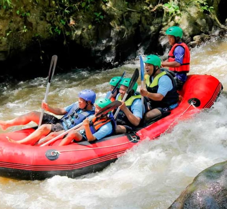 A group of people wearing helmets white water rafting in a red boat on a tropical river.
