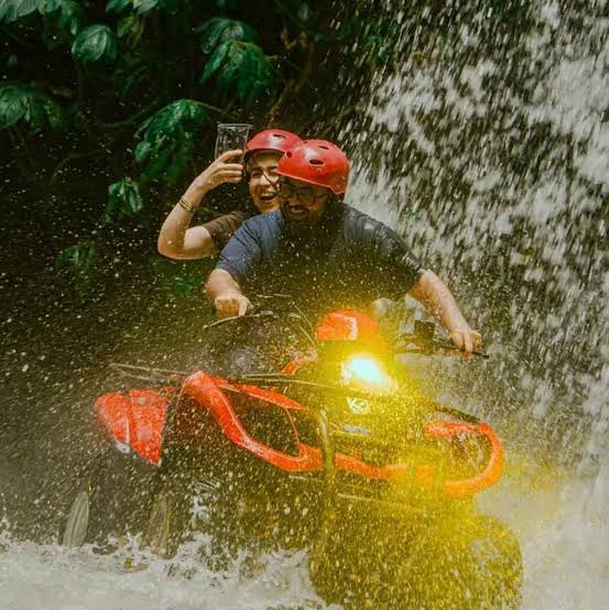 Two people wearing helmets ride a red ATV through a tropical waterfall during an outdoor adventure tour.