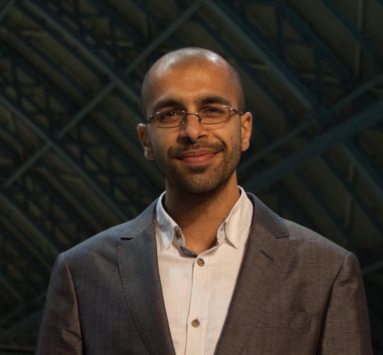 A smiling professional man with glasses and a grey blazer in a modern office setting.