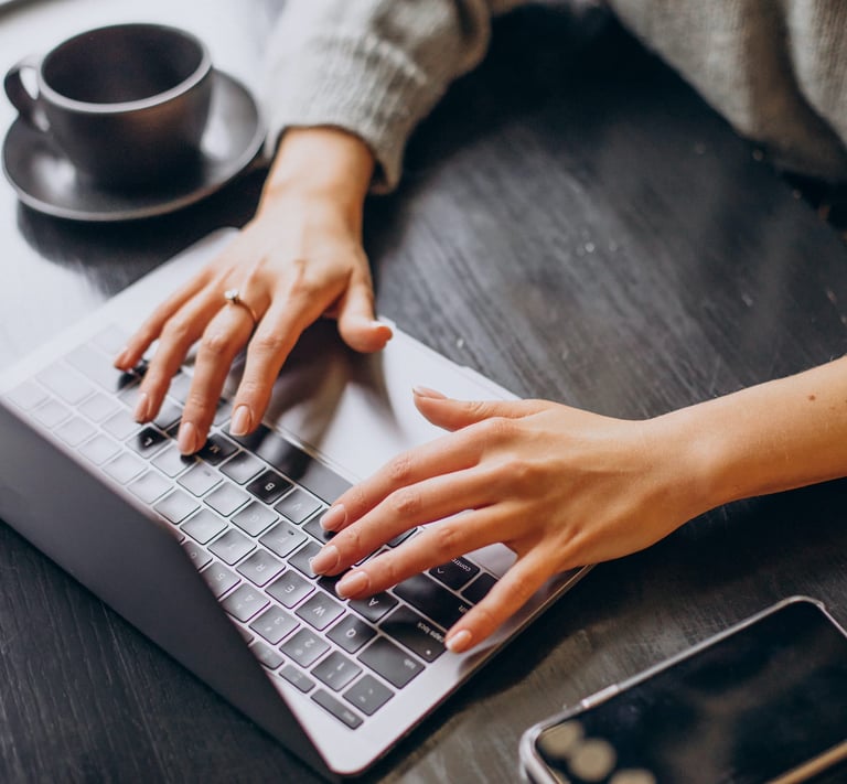 Een persoon die aan een tafel zit met een laptop en een kopje koffie, voor de contactpagina