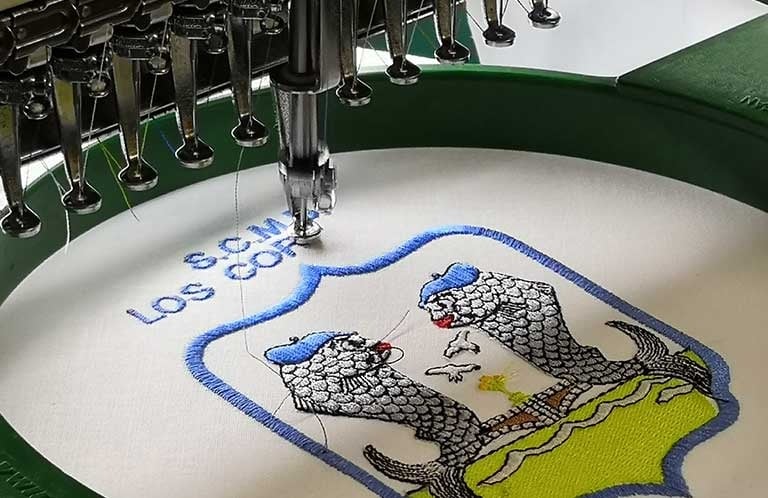 Industrial embroidery machine stitching a custom logo crest with blue and green thread.