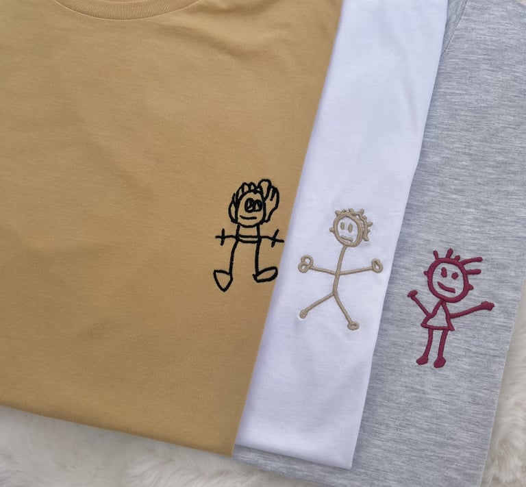 Custom embroidered t-shirts featuring personalized children's stick figure drawings in mustard, white, and grey.