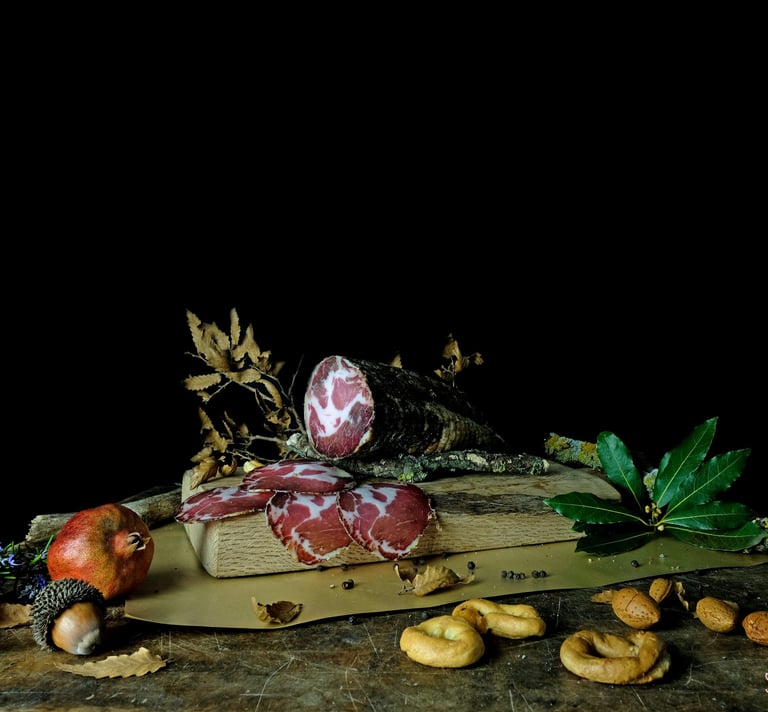 Slices of cured capocollo ham on a rustic wooden board with taralli, almonds, and pomegranate.
