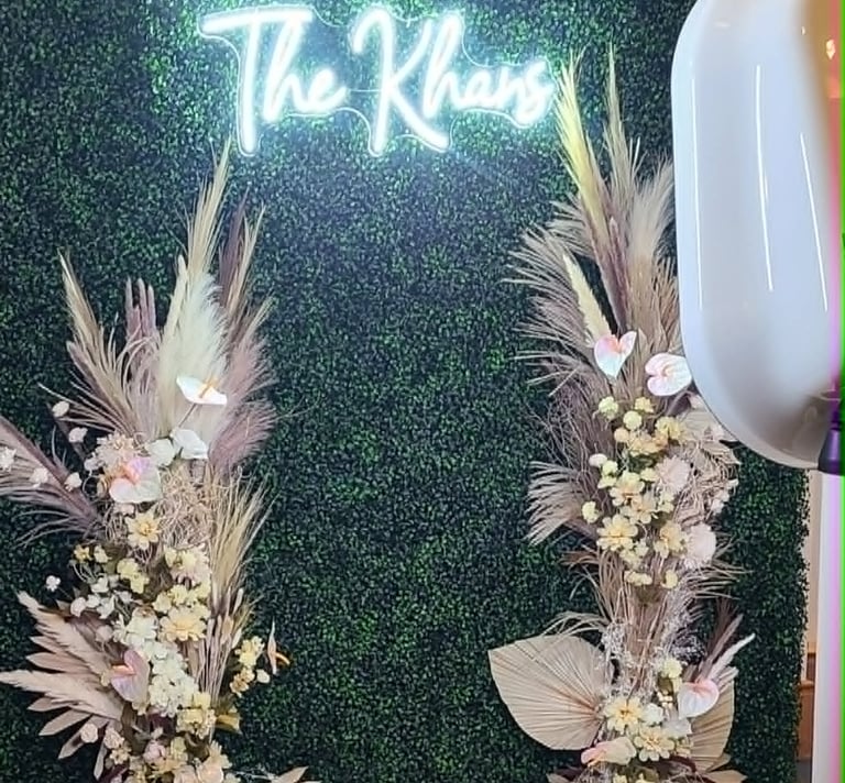 A wedding photo booth backdrop with a neon sign, green boxwood wall, and dried floral arrangements.