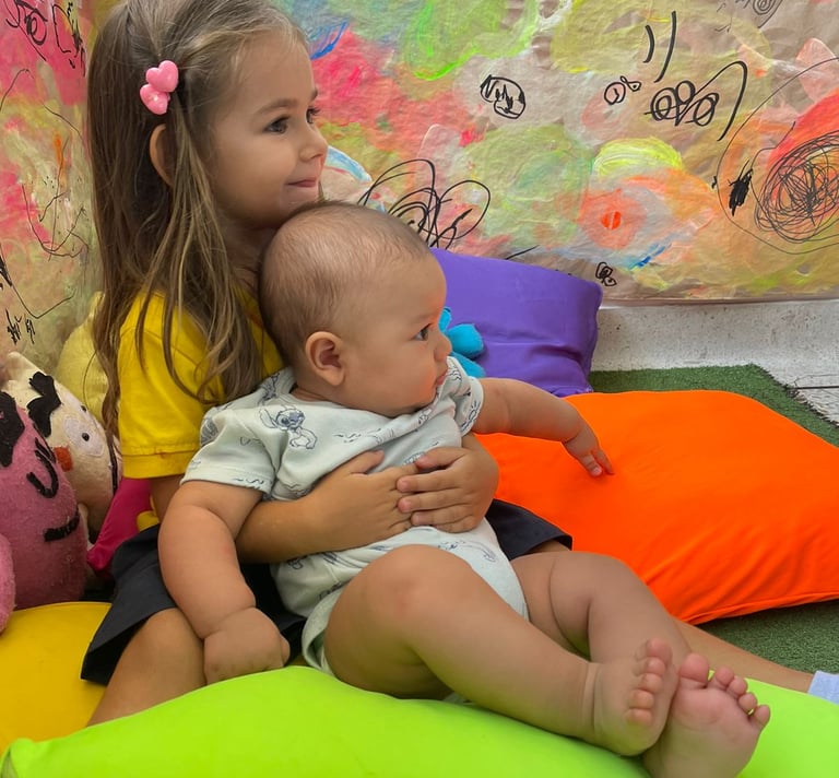 Niña abrazando bebe sentados entre cojines de colores vibrantes en Ukelele Jardín Infantil