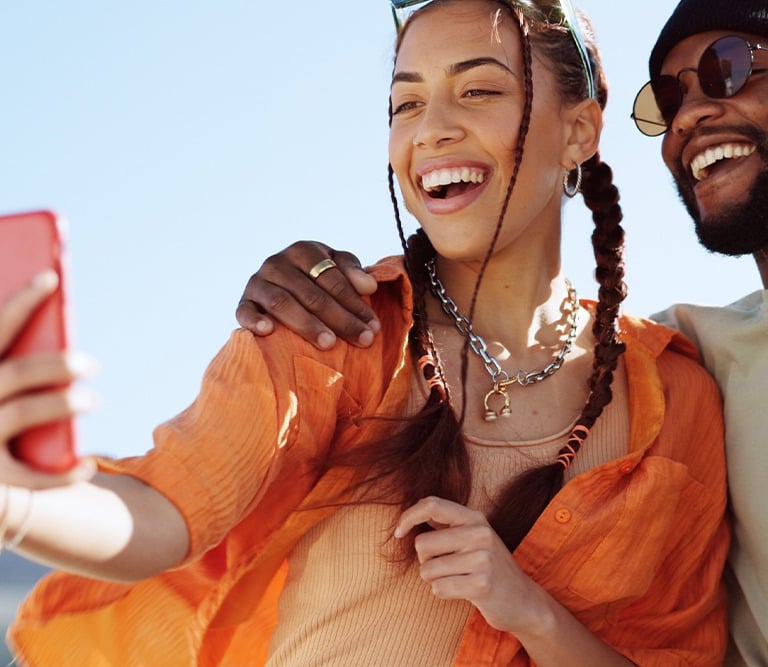 A smiling young couple takes a selfie on a red smartphone with mountains in the background.
