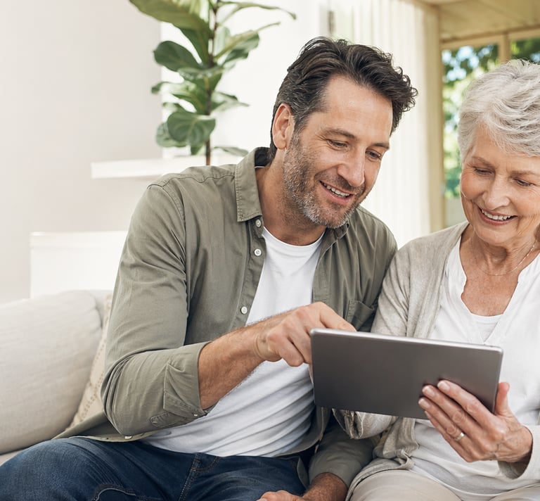 A smiling adult son and senior mother use a digital tablet together on a living room sofa.