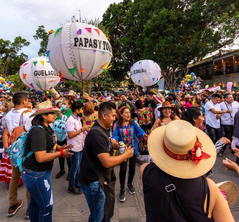 Gente bailando en calenda de Guelaguetza en Oaxaca con globos gigantes y música de fondo
