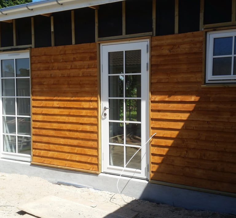 Moderne havebygning under opførelse med vandret træbeklædning og hvide glasdøre.