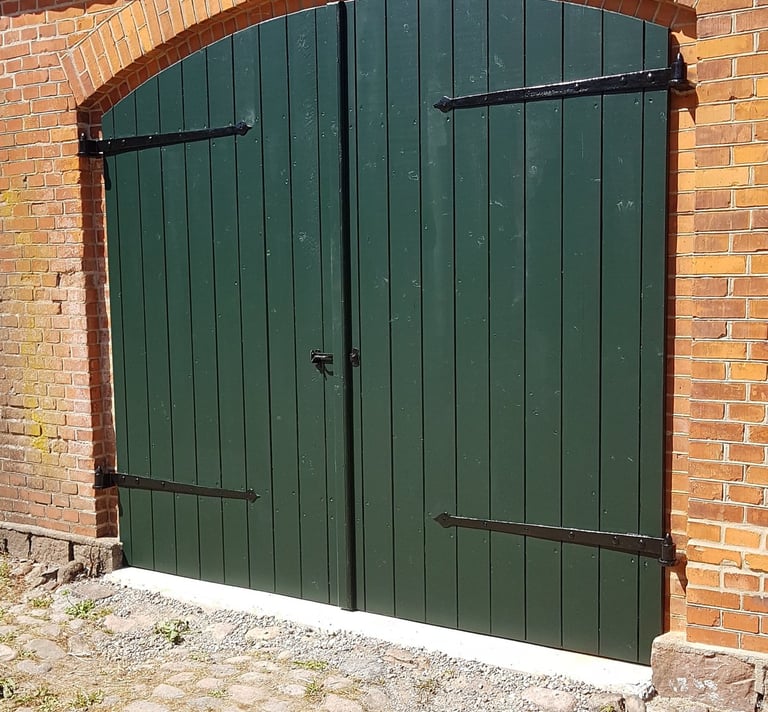 Mørkegrønne buede træporte med sorte metalhængsler, indsat i en rustik rød murstensvæg.