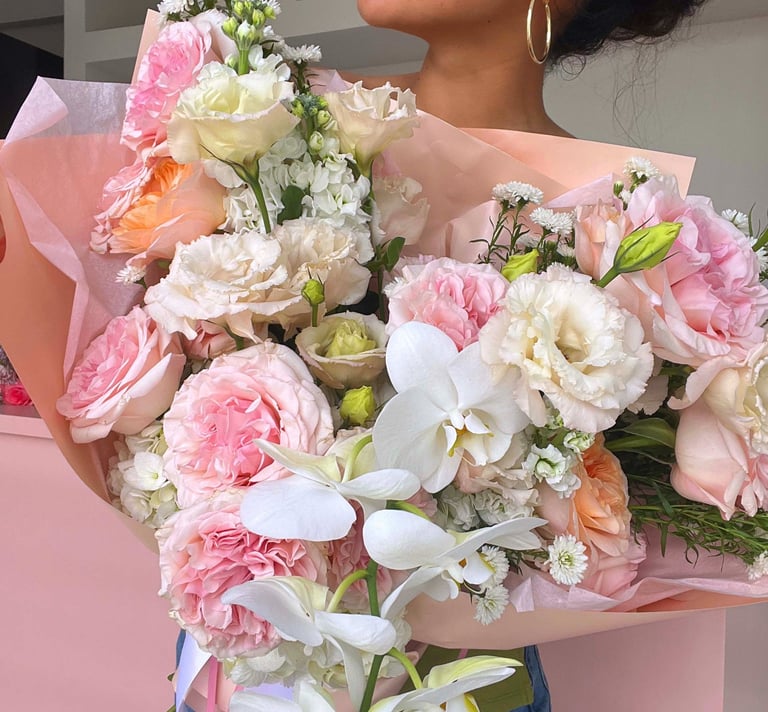 A woman holding a large floral bouquet featuring pink roses, white orchids, and lisianthus flowers.