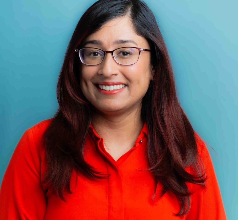 A smiling woman in glasses stands against a blue background, wearing a vibrant red shirt and a friendly demeanor.