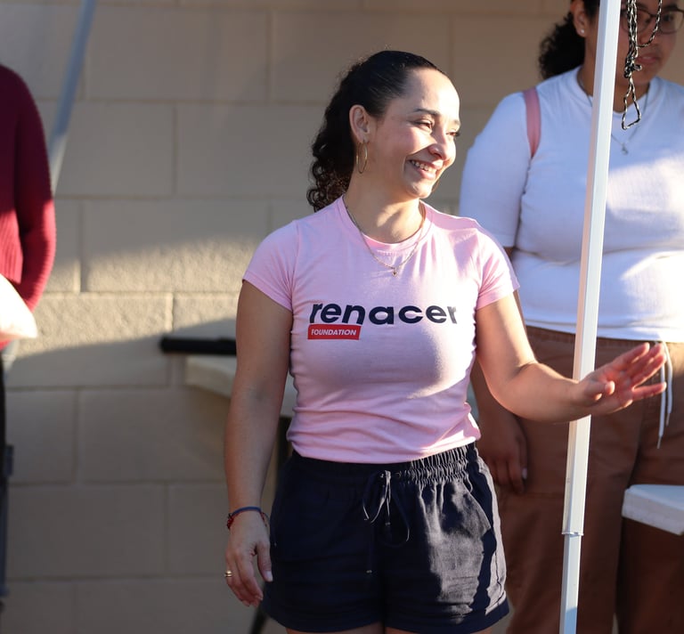 A smiling woman wearing a pink Renacer Foundation t-shirt volunteering at a community outreach event.