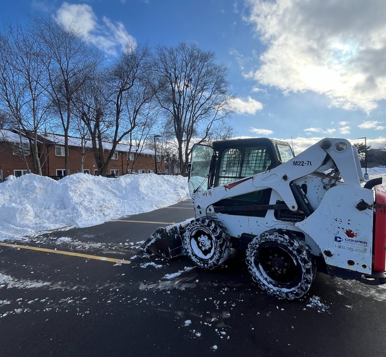 White Bobcat skid steer loader clearing a snow-covered commercial lot near residential buildings.