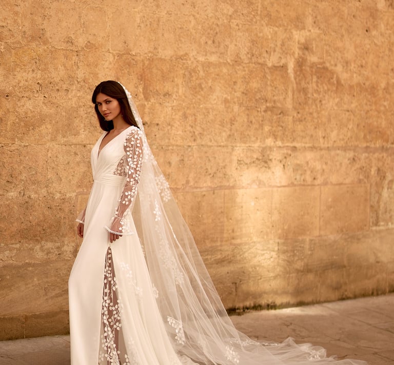 Elegant bride wearing a long sleeve lace wedding dress with a floor-length floral veil against a stone wall.