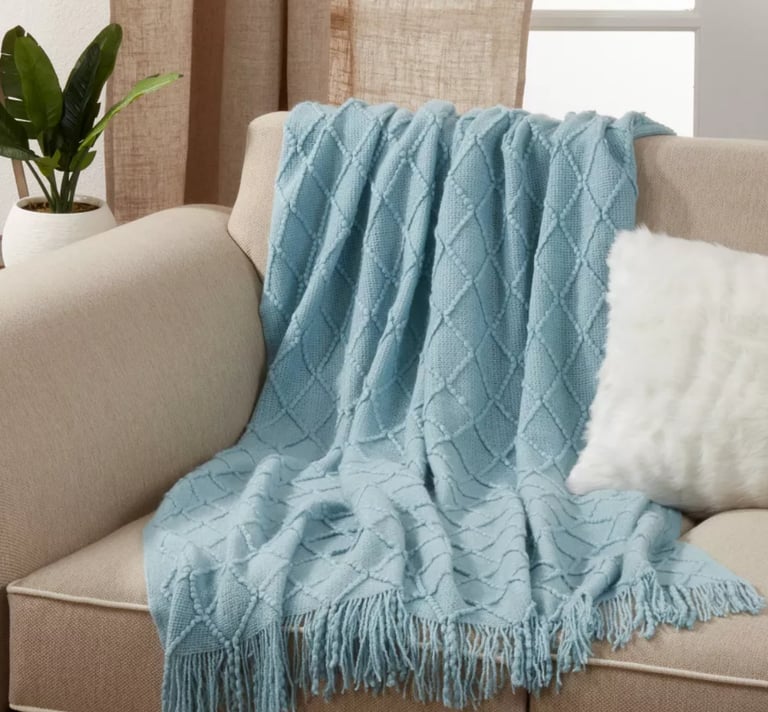 Light blue diamond pattern knitted throw blanket with fringe draped on a beige sofa.