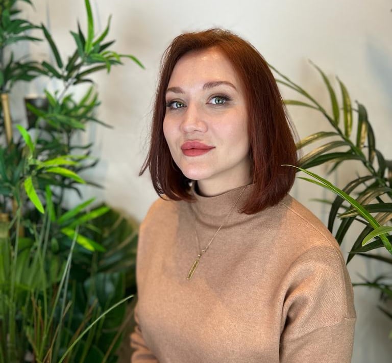 Woman with a professional auburn bob haircut wearing a tan turtleneck sweater among green plants.
