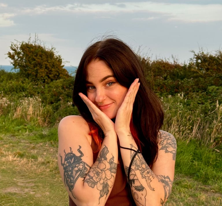 Smiling woman with dark hair and floral arm tattoos posing outdoors in nature at sunset.