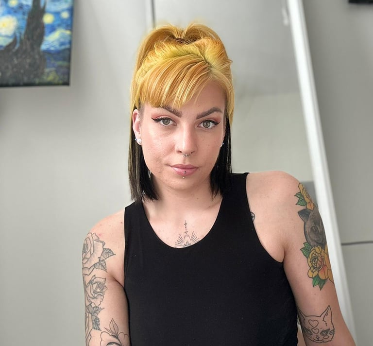 A woman with yellow and black split-dye hair and arm tattoos wearing a black tank top.