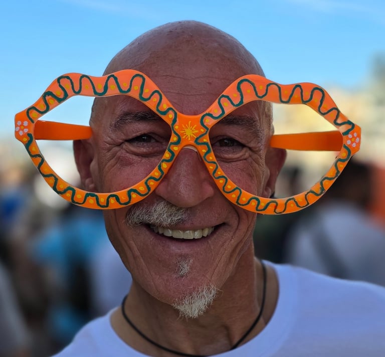 Older man smiles, wearing big colorful lips-shaped glasses outdoors, blue sky above and a blurred crowd behind him.