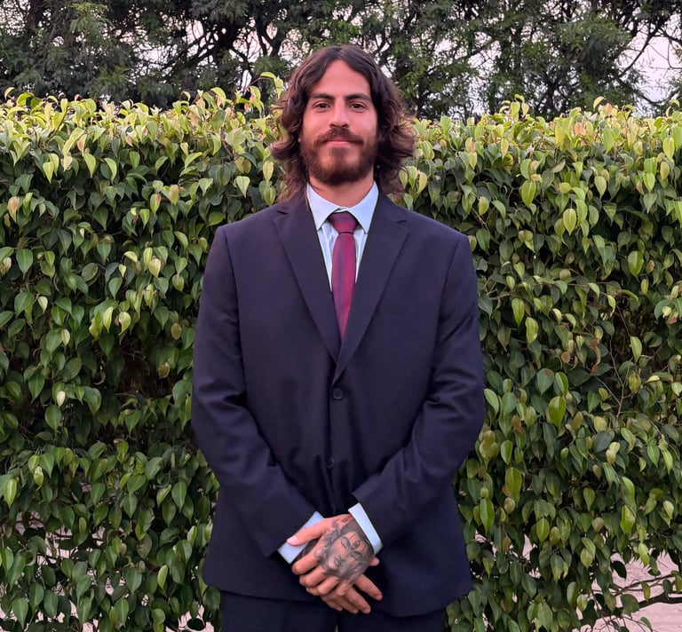 A smiling man with long wavy hair and a beard wearing a formal suit and tie outdoors.