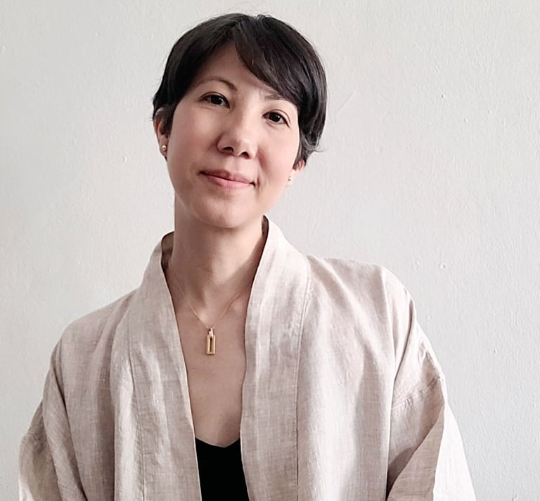 A smiling woman with short black hair wearing a beige linen cardigan and gold necklace against a white wall.