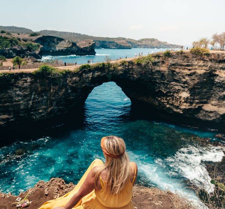 A blonde woman in a yellow dress sits overlooking Broken Beach in Nusa Penida, Bali.
