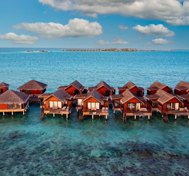Row of overwater bungalows built on stilts above a turquoise ocean, with a distant island visible un