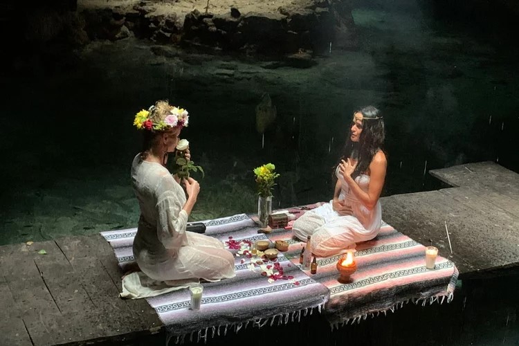 Two women seated on colorful woven blankets performing a candle-lit ritual inside a cenote in Tulum,