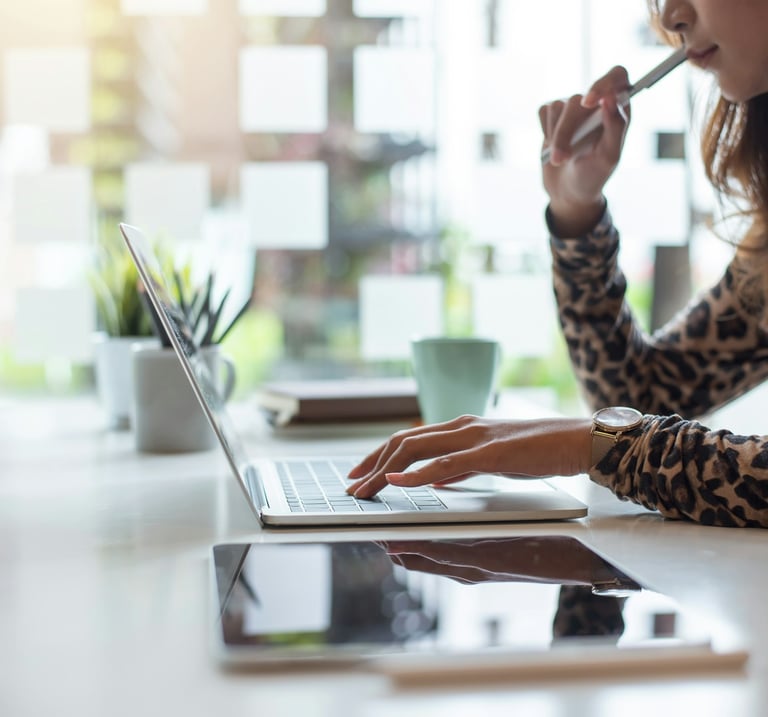 Professional woman holding a pen near her mouth, working on a laptop on a desk with a digital tablet, plant, and coffee cup.