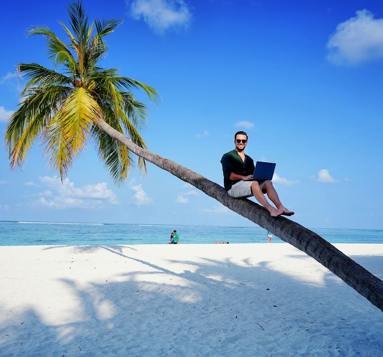 A digital nomad working on a laptop while sitting on a leaning palm tree at a tropical beach.