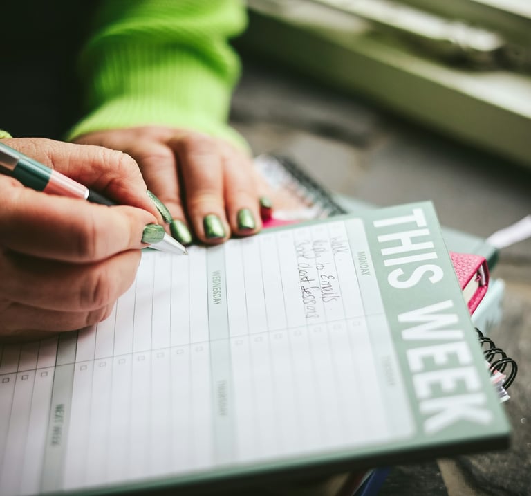 A person writing in a green weekly planner pad to organize their schedule and tasks.