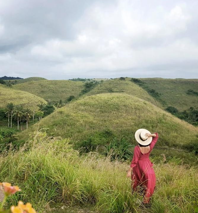 A woman in a red dress and sun hat overlooks lush green rolling hills and tropical landscape.