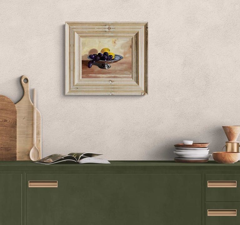 Framed still life oil painting of grapes and lemons hanging above a modern olive green kitchen cabinet.
