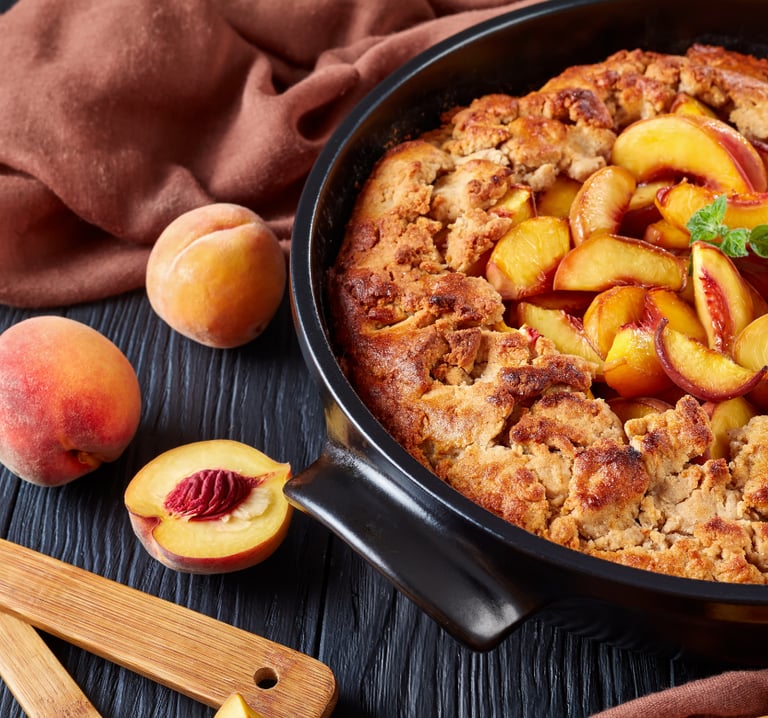 Freshly baked warm peach cobbler in a black skillet with sliced fresh peaches on a dark wooden background.