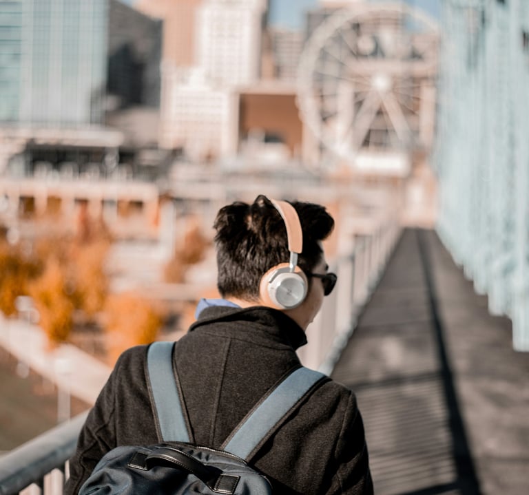A man wearing wireless headphones and a backpack walks across a bridge with a city skyline and ferris wheel.