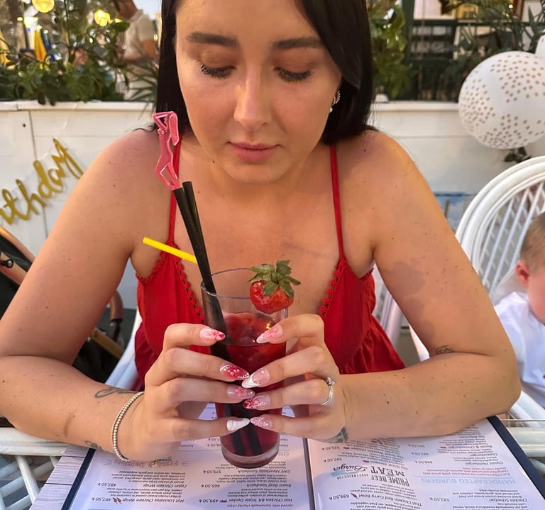 Young lady in red dress on vacation, holding a cocktail in a restaurant brilliantly displaying beautiful nails by Little Lift