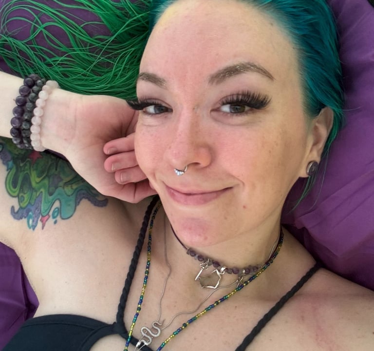 Anointistry creator, woman with green hair, crystal jewelry smiling warmly laying on a purple bed 