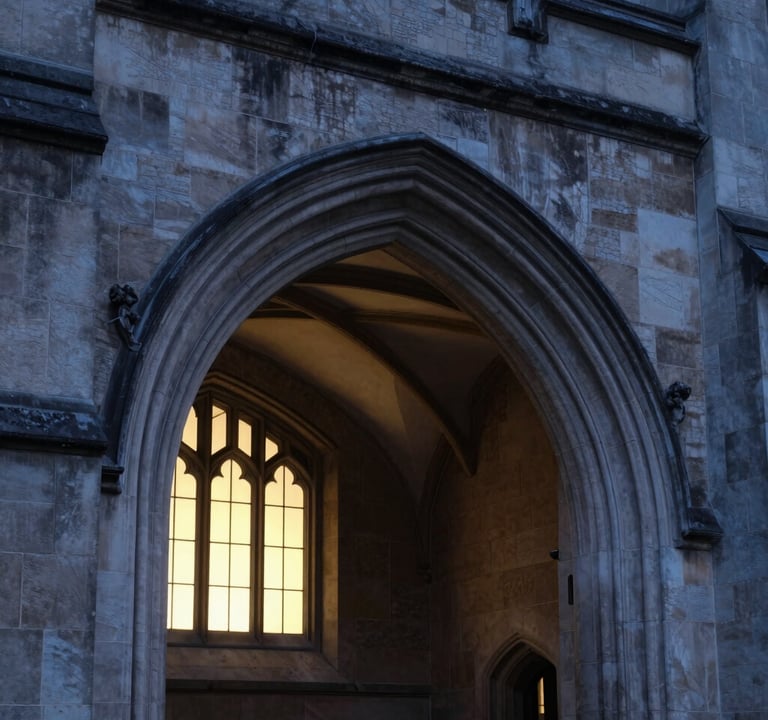 An architectural detail of a Gothic stone archway at a prestigious university during the blue hour. The textures of the charcoal-colored stone are sharp and detailed, with cream light spilling from a window in the background.
