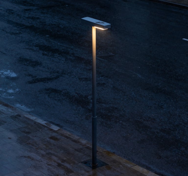 A minimalistic street life photograph during a rainy evening. A single modern street lamp illuminates wet, dark navy pavement. The composition is clean and empty, emphasizing solitude and elegance. International / Western urban environment.