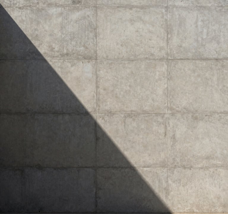 A striking architectural photograph of a minimalist concrete facade in the Middle Eastern / Gulf region. The sun creates a sharp, diagonal shadow across the surface, highlighting the rich textures. Minimal, expensive, and modern.