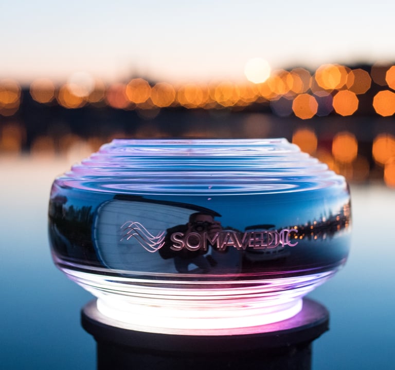 Glowing Somavedic EMF mitigation device outdoors at sunset with bokeh city lights background.