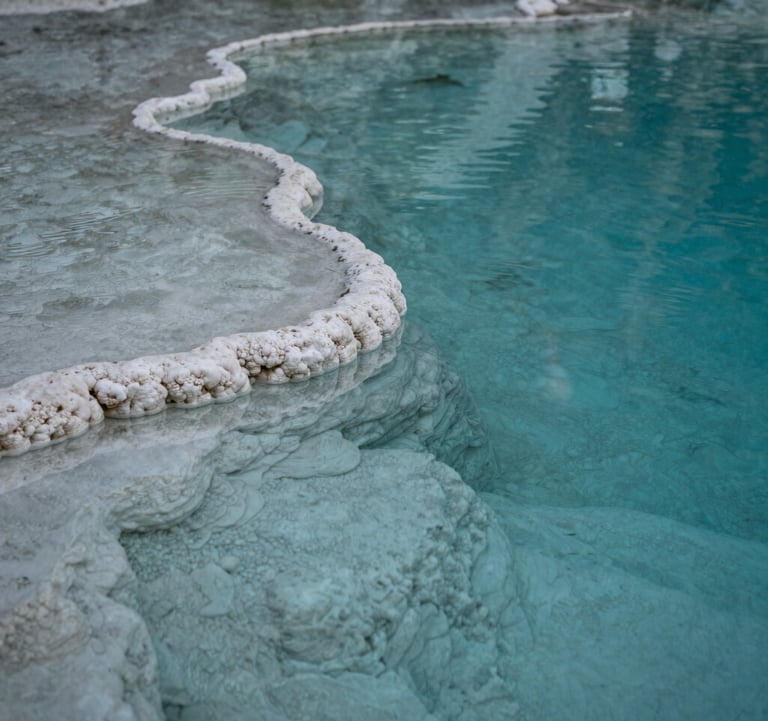 A close-up photograph of a natural mineral pool at Hierve el Agua, with crystal clear turquoise water reflecting a soft white stone edge. The lighting is soft and contemplative, highlighting the textures of the mineral deposits.