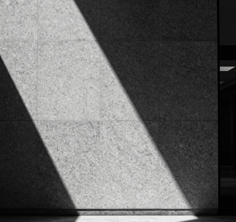 A minimalist, high-contrast black and white photograph of a single shaft of light illuminating a granite texture on a wall in a North American / International Business lobby. Sharp shadows, intellectual precision.