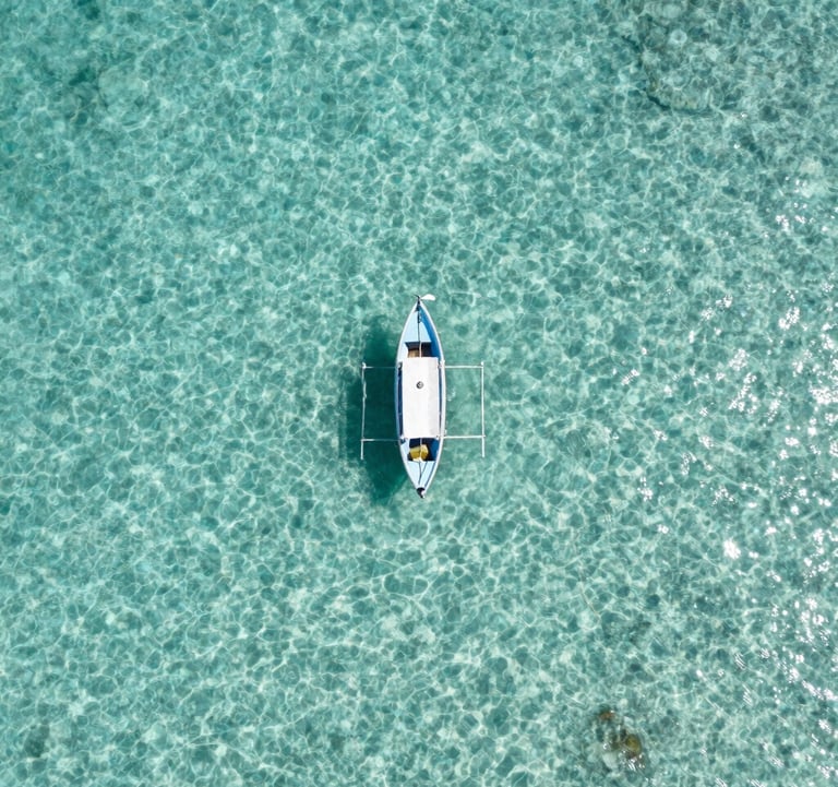 An aerial photography shot of a single small boat floating on crystal clear, light cyan water in the South American Caribbean. The composition is clean and minimalist, showcasing the depth and clarity of the ocean.