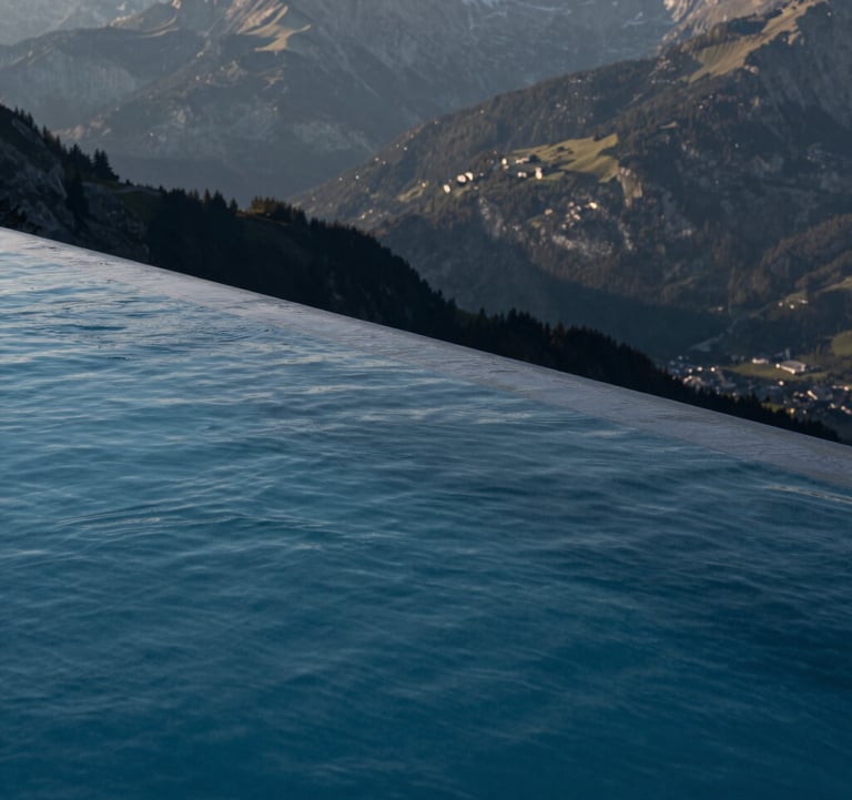 Detail of a luxury infinity pool overlooking the Swiss mountains, sharp architectural edges, deep night blue water, morning sun, high-end photography for an architecture magazine.