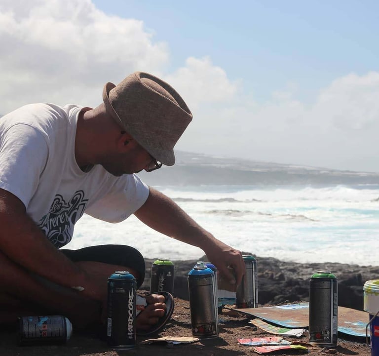 Artist in a fedora hat preparing spray paint cans for a mural on a rocky coastline.