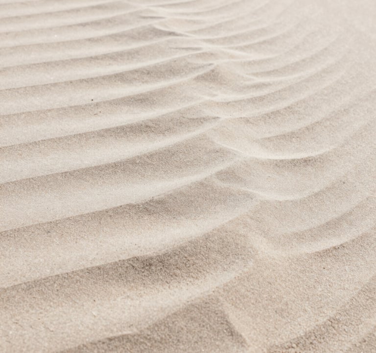 A close-up shot of wind-blown sand forming patterns that mimic ocean waves. The colors range from deep #DDCAB8 to light #FAF8F5, emphasizing texture and natural design.