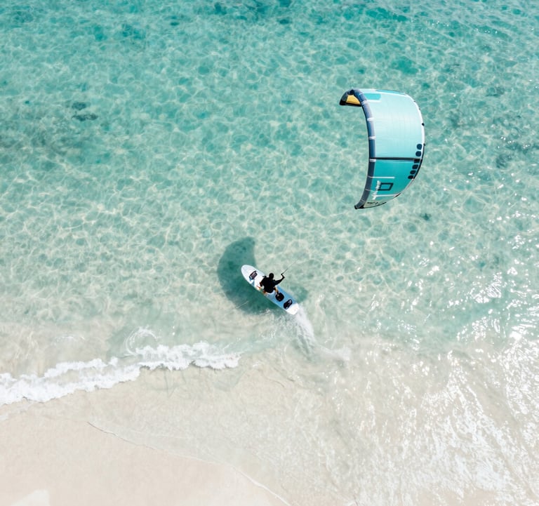 An artistic, top-down view of a kitesurfing board partially submerged in crystal clear turquoise water, casting a soft shadow on the white sand #FAF8F5 below. The composition is minimal and highlights the movement and energy of the ocean.